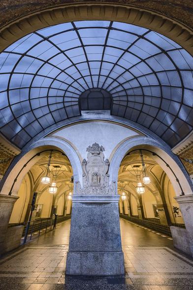 Vertical, symmetrical, wide-angle view of the entrance hall to the platform at Heidelberger Platz U-Bahn station in Berlin. A circular blue stainglass dome covers the hall, the light coming down from it covering in a light blue shade the grey stones, the main pillar holding the dome, including its adornement. By contrast, the cathedral-style platform behind is baved in yellow lighting from the lamps hanging from the ceiling.