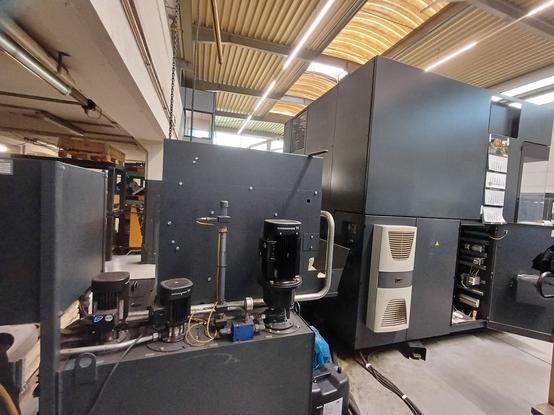 A wider view of an industrial CNC machine setup in a factory. Large black cabinets house electrical and automation components, while pumps and cooling systems are visible in the foreground. The factory ceiling with lighting and ventilation systems can be seen.