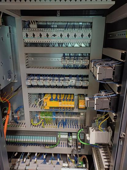 An open electrical control cabinet with various relays, circuit breakers, and industrial automation components. Cables are neatly routed to different terminals, and several Siemens PLC modules are installed.