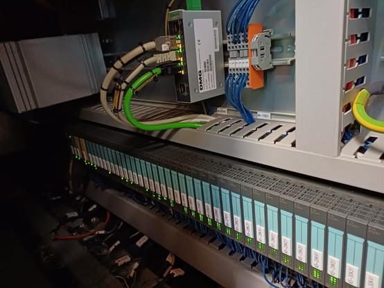 A close-up of a control panel with multiple Siemens Simatic I/O modules. Thick industrial cables are routed through cable trays and connected to different modules.