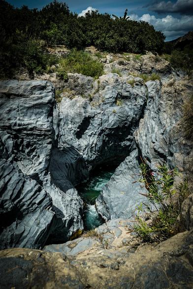 The image depicts a rocky gorge with a river flowing through it. The gorge is formed by dark, layered rock with a rough texture, showcasing a mix of gray and black hues. The river, visible at the bottom of the gorge, has a turquoise color, indicating clear water. The surrounding area is covered with green vegetation, including bushes and trees, which contrast with the rocky terrain. The sky above is partly cloudy, with patches of blue visible. The scene is illuminated by natural light, highlighting the ruggedness of the rocks and the vibrant greenery.

Provided by @altbot, generated privately and locally using Ovis2-8B