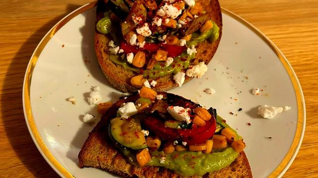 Two slices of toasted bread with guacamole and grilled vegetables, vegan white cheese and pul biber on a white plate on a wooden surface.