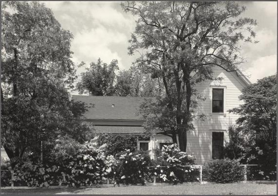 The image depicts a black and white photograph of an old, two-story house nestled among trees with lush foliage. The architecture suggests it may be from the early to mid-19th century or possibly earlier. There's no visible activity around the property which contributes to its tranquil appearance. In front of the home is what appears to be a well-maintained garden filled with an array of flowering shrubs, likely indicative of springtime when such blooms are most vivid.

The sky above shows some cloud coverage but overall it seems like a bright day as indicated by the shadows cast on and around the house. The lack of any modern elements or cars suggests this photograph might be from several decades ago. There's nothing that indicates current occupancy, hinting at abandonment or a deliberate choice to maintain its rustic charm.

Given the source information provided (14A. Home Place), it is plausible that this image was taken as part of an ongoing documentation project focused on various homes and their settings during mid-20th century America. The photographer Dorothea Lange, known for her work in social documentary photography especially during times of great upheaval such as the Great Depression, further adds context to why these particular structures might have been subjects of interest.

The reference number indicates that this photograph is part of a larger collection or series and it could be used to identify specif [...]