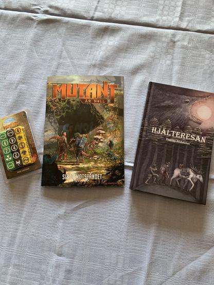 Ett fotografi av tre nyinköpta artiklar som ligger upplagda på en blå duk: ett blister med gröna, gula och svarta tärningar till rollspelet Mutant År Noll; en äventyrsbok till rollspelet Mutant År Noll med titeln ”Sista Moståndet”; och en hårdpärmsbok med titeln ”Hjälteresan” av författaren Tommy Bildström.