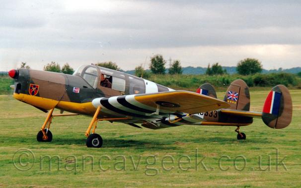 Side view of a small, single propellor-engined light aircraft with 3 tail-fins, an olive green and brown camouflage scheme on the top surfaces, and a yellow belly and all lower surfaces, taxiing from right to left on grass.
There are "D-Day Stripes" (white-black-white-black-white bands) around the wings just outboard of the fixed undercarriage and rear fuselage.
There is a red shield with a yellow outline and a large black cross in the centre with crossed swords on the planes nose, red, white, and blue roundels visible under the wings, a red, white, and blue fin flash on the leading edge of the inner and outer faces of the outer tail-fins, and a British flag over 4 gold stars on the central fin.
A man can be seen sitting in the pilots seat through an open cockpit window.
Short green grass fills the foreground, leading up to bushes and trees at the airfield perimeter in the distance, under cloudy grey skies.