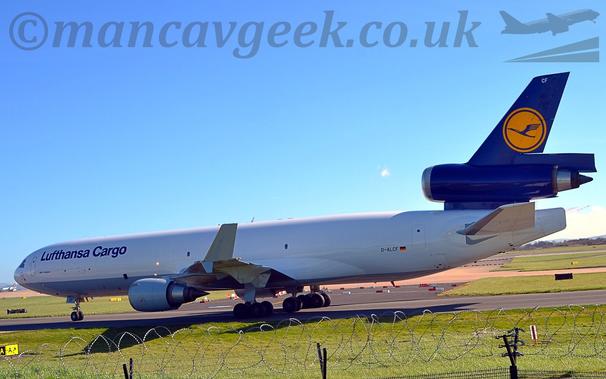 Side view of a white, 3 engined jet freighter taxinng from right to left.
The plane has a grey belly and engine pods slung under the wing, with dark blue "Lufthansa Cargo" titles on the upper forward fuselage.
The tail and centre engine are a dark blue, with a large yellow circle containing a stylised blue flying bird halfway up the tail-fin.
The upturned wingtips are white, as are the smaller downturned tip.
Green grass fills the foreground, behind rolls of barbed wire on top of a black metal fence.
Under the plane in the background, patches of grass separate taxiways and runways.
Above, pale blue sky fills the rest of the frame.