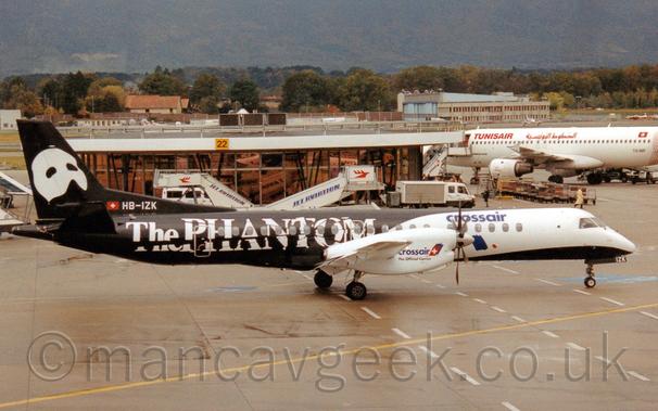 Side view of a black and white, twin propellor-engined airliner taxiing from left to right.
The planes rear fuselage and tail is black, with fractured white "The Phantom" titles on the side of the body, and a white mask designed to be worn over the upper part of the face and across the bridge of the nose on the tail.
The forward fuselage and wing-mounted engine pods are white, with blue "CrossAir" titles on the upper body and on the engine pods
The registration "HB-IZK" is on the bottom of the tail in white, next to s small red Swiss flag.
Grey concrete apron covered with yellow lines and white dashed lines fills the foreground, with a circular, heavily glazed terminal building in the background.
A larger, white jet airliner with red "TunisAir" titles is parked next to this terminal building.
Trees in the distance run off towards the horizon, leading to the lower reaches of a tree-covered mountain rising into the sky.