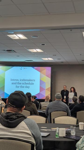 A room of people listening to the summit organizers welcome everyone and run down the schedule for the day. A big projected slide says, "Intros, icebreakers and the schedule for the day."