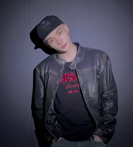 Hyunjin facing the camera with his hands in his pockets and his head tilted to one side. He's wearing a black cap with a black cross on it