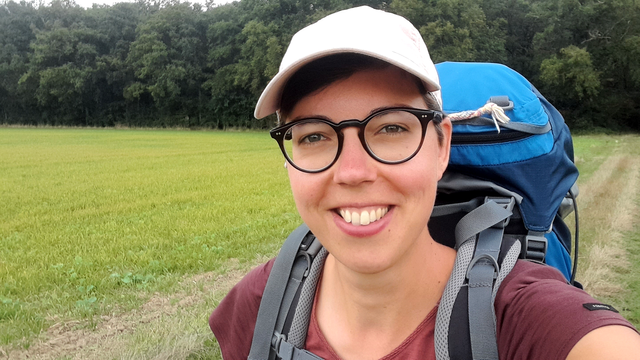 A selfie of a white person wearing a pink cap and smiling. I am wearing a short-sleeved t-shirt and a large blue backpack. In the background is grass and further away a line of trees.