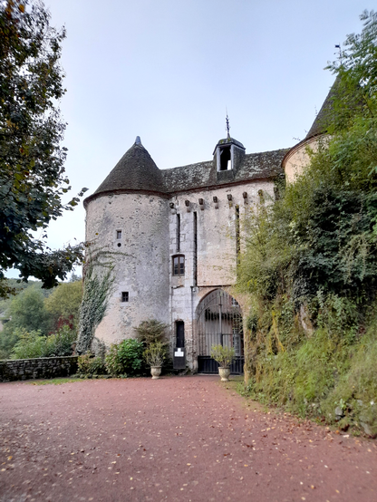 A courtyard in front of a small castle, which is partly obscured by bushes to the right. The gate is closed.