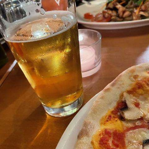 A mouth-watering close-up of a cold beer in a frosted glass next to a pizza on a white plate. The background shows a candle and another dish, suggesting a relaxed dining atmosphere, likely in a restaurant or pizzeria.