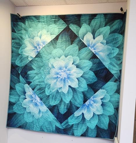 A quilt hanging on a wall, in a variety of shades from green-blues to blues to black. The center is a square turned to an angle, making a diamond shape. In that diamond is a large green-blue flower. On each outside edge of the diamond is a quarter-reflection of the inside flower design in the same colors. (Design and needlework by Laurie Kay Janda.)