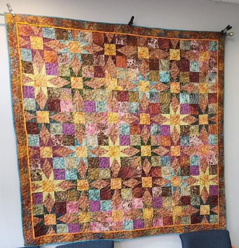A large quilt hanging on a wall, in shades of brown, yellow, and just a tiny bit of pink and baby blue. Inside the golden-brown border is a repeating pattern of "log cabin" star designs. Often times, the colors are muted and blend so perfectly with the ones next to them that it is hard to distinguish the star patterns in every quilt block. This gives a feeling of depth to the stars, as if they are floating in a celestial sky of fabrics. (Design and needlework by Laurie Kay Janda.)