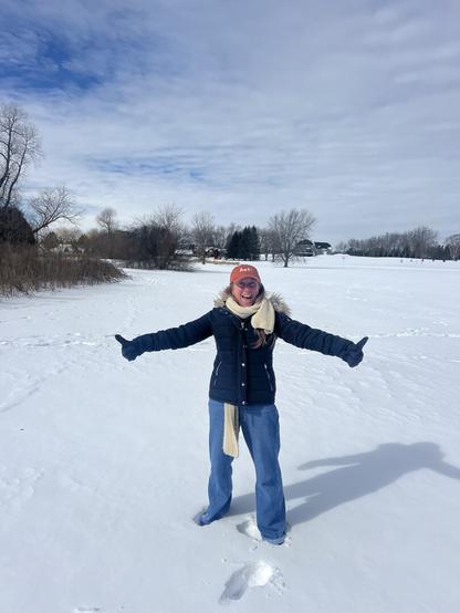 woman standing, all bundled up, in a very snowy landscape. Her arms are outstretched with thumbs up- she is about to make a snow angel. Smiling... and blue sky above.