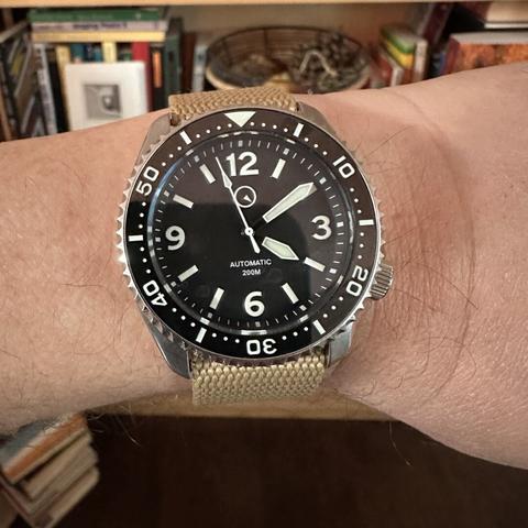 Watch with a brushed stainless steel case, 4:00 crown, black dial with numeric indices at 12, 3 , 9, and 6, baton indices elsewhere, white lume-filled sword hands, and black ceramic dive bezel with white numerals and markers, on a khaki fabric strap.