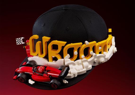 Black cap with yellow LEGO "VROOM" text and a small red LEGO race car attached, set against a dark red background.