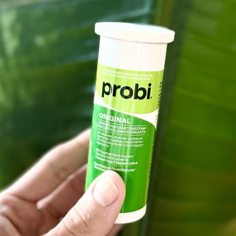 A hand holding a green cylindrical container labeled "Probi Original," which contains vegetarian capsules with patented bacterial strains. The background features blurred green leaves.