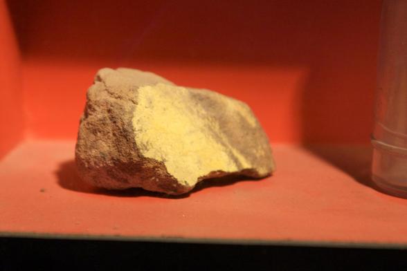 Close-up photograph of a piece of raw uranium on an orange background. From the New Mexico Mining Museum. By Marcin Wichary, under a free flickr license: https://bit.ly/3R1e6RB.