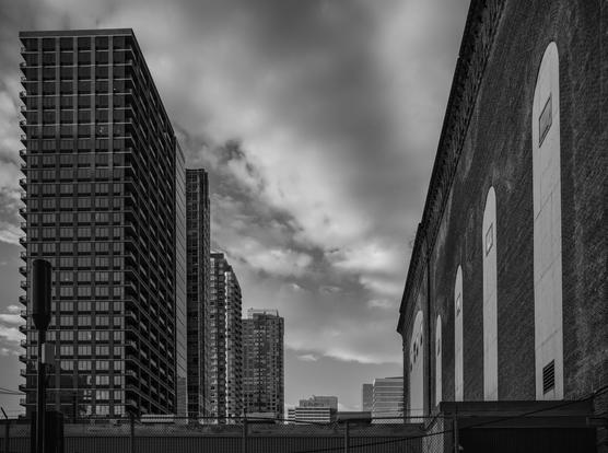 A monochrome photo looking down a corridor between rows of buildings.  There are recent high-rise towers on the left, and an earlier brick industrial building on the right.  Where sky is visible, the clouds are dramatic.