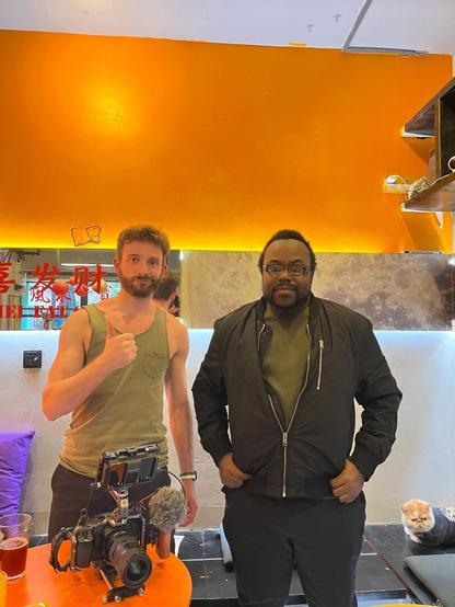 Two men stand behind a table in an orange room. One gives a thumbs up. A camera sits on the table and there is a cat in the corner