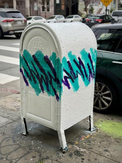 A US postal box that has been painted all white, with teal and purple zigzags running along the middle of it. This was done by a street artist and it resembles a disposable cup by Dixie Cups.