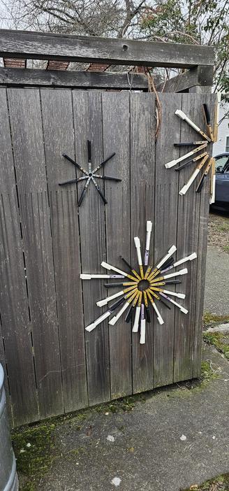 A fence made of weathered cedar is decorated with stars built with industrial leftovers, which i can't identify. One star in the upper left is made with black metallic strips joined with what look like screwed in shelving strips . A large star on the bottom has rays of white metallic parts ( computer? drawer pieces?) and black strips joined by yellow wooden strips. One that wraps around the corner at the top right is a copy of this last one. 

I don't know what the various strips are from!