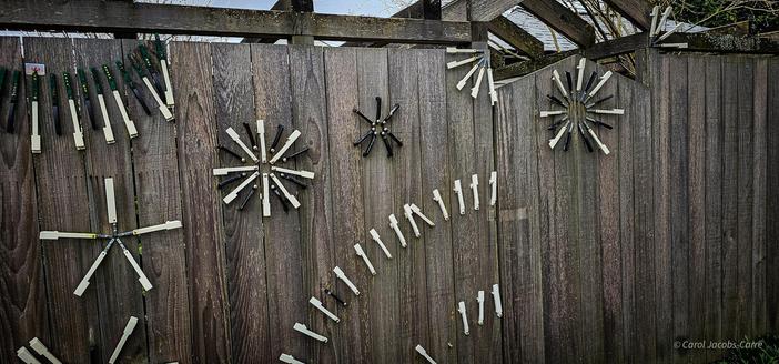 A street-facing continuation of the weathered cedar fence has stars as described before, and three curves framing the stars one at the top made of white and black strips set closely spaced side by side, and two at the bottom, made of two series of white strips set closely spaced side by side.
It looks very modernist and hand made. There is a gate towards the right side with a black and white star at the top.