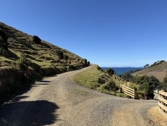 A winding gravel road curves through lush green hills overlooking a blue ocean under a clear sky. Nearby vegetation includes low shrubs and trees, with a wooden fence visible in the foreground. Mind the ocean in the background.
