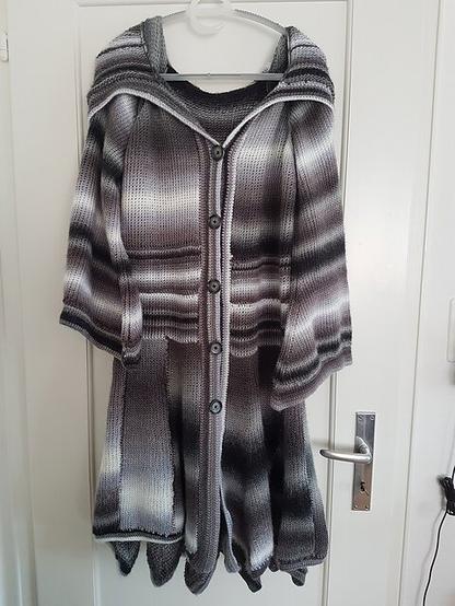 A long, flowing cardigan crafted with Tunisian crochet hangs on a white door. The cardigan features a hood and a gradient pattern transitioning through shades of gray, black, and white. Its design includes a button-up front with five buttons, pockets, and a loose, draping style at the bottom