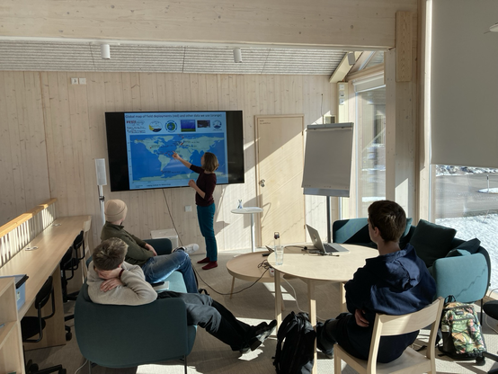 A scientist exlains something in front of a screen. Three Phd students listening.