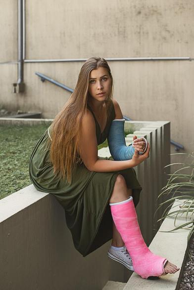 Eine junge Frau sitzt mit Gehgips und Gipsarm auf einem Mauervorsprung in einer urbanen, brutalistisch angehauchten Umgebung.