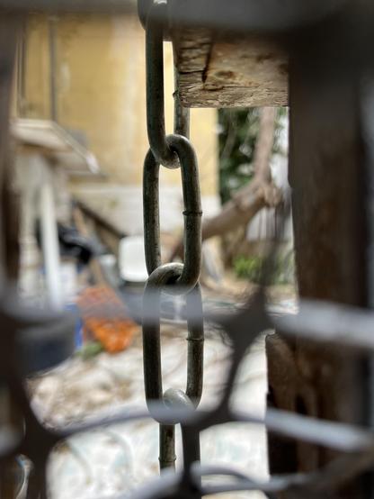 Through a gap in a metal gate, the out of focus courtyard of an abandoned house. Sharp in the centre of the image, the chain holding the gate shut.