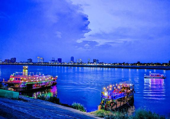 Photo shows a deep blue evening just after sunset with two party/evening dinner river trip boats just about to leave with guests. The boats are well and partylike deorated with lights. In the background is the river Tonle Sap with a couple smaller boats, and the houses on the other river side. In the background the evening sky is split in two, a lighter dark sky to the east and deep dark stormy rainy clouds. Where the dark clouds meet the lighter side, the clouds forms a profile reminiscent of a elder gentleman.