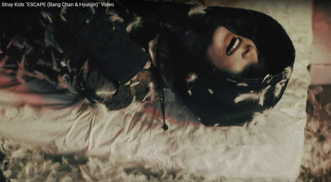 Hyunjin lying on his back on the grotty mattress. He's wearing a dark hoodie that obscures his eyes. His mouth is open and happy. There are feathers everywhere like a pillow exploded.