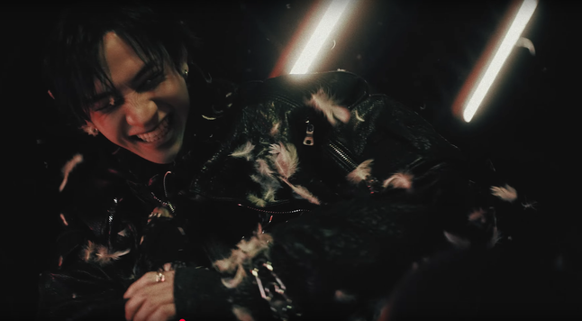 Feather-covered Bang Chan grinning maniacally, as seen from the perspective of a person that he's just thrown down onto a mattress