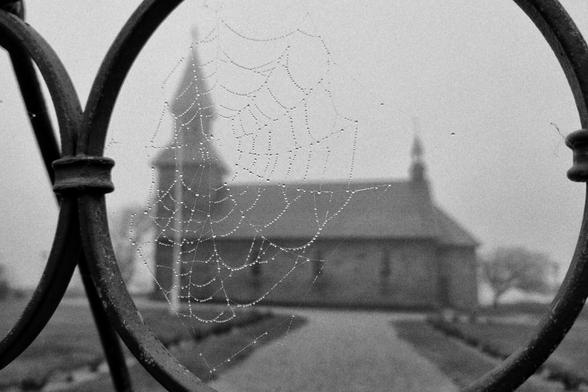 Black and white shot of a church in Gudhjem on Bornholm. The church is framed by an iron gate in the very near forground on which a spiders Web hangs heavy with water droplets from seamist.