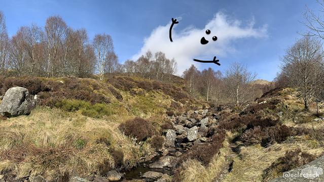 A photo of a beautiful mountainside with a small rocky stream running down it. There are large lichen covered boulders, patches of still dark brown heather, mossy grass and bare, just budding trees. In the distance is a mountain peak, with a blue sky above. There is one cloud poking up from behind the mountain. It has a smiling face and upturned arms, greeting you to this lovely place.