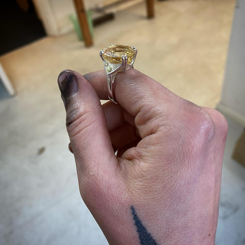 A hand with dirty blackened fingers is holding a shiny silver ring. The ring is set with a bright yellow centre stone, it is held in place by four double claws. You can see an out of focus workshop floor in the background