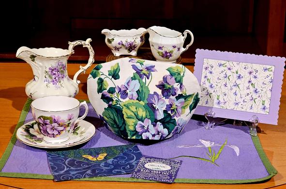 Tea tableau in purple, green, and lavender with linens, paper, and several vintage china pieces patterned in violets. There is a placemat with an embroidered iris, and St Dalfour Earl Grey black tea.