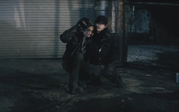Hyunjin and Bang Chan looking a little scared as they crouch together, on the run from something. Chan is covering Hyunjin's mouth with his hand