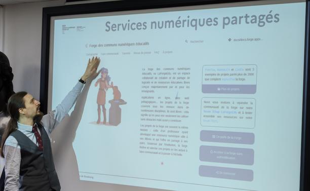 Un homme de profil présente la page d'accueil de La forge des communs numériques éducatifs sur un écran de vidéoprojection. De la main gauche il montre l'accès à la cartographie des ressources de la forge des communs numériques éducatifs.