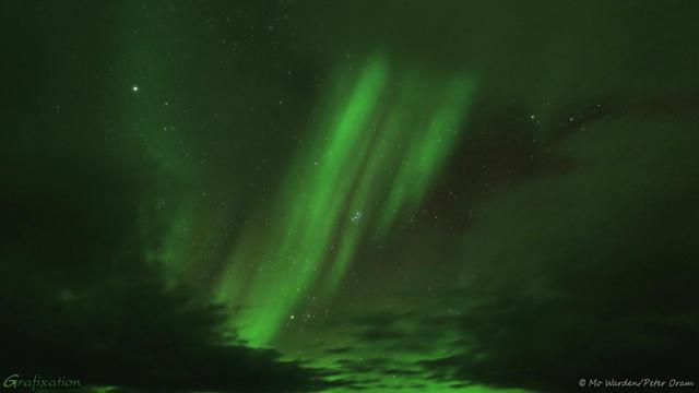 A colour photo of the night sky, with heavy cloud around three sides except for a gap at the bottom. Many stars are visible, including the Pleiades. Through the centre of the shot is a bright green band of diagonal aurora, streaked and striated.