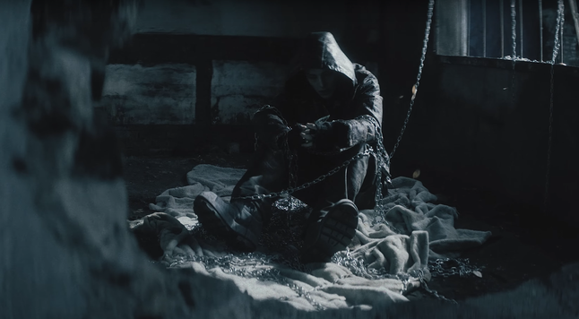 Hyunjin sitting on the floor in a dark, blue-tinged room. There's a rumpled rug underneath him and chains draped across and around him.