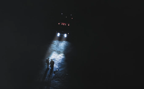 Wide shot of two boys holding hands in the dark, lit by blue lights on the front of a large vehicle. There are red lights on the vehicle, higher up, but they don't illuminate anything.