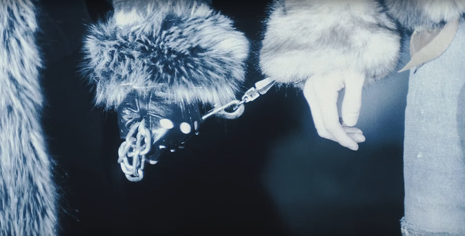 Close shot of the hands of two boys who are standing next to each other, wearing fur coats. One has a length of chain attached to his wrist. The other is wearing a leather glove with cut-outs, and has a firm grip on the loose end of the chain.