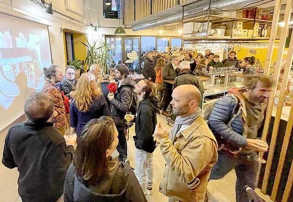 Photo prise au début de la soirée d’anniversaire du « 8 Fablab », au bar coopératif « Le central vapeur ». Au premier plan, quelques personnes, debout avec un verre à la main, discutent devant des images vidéo-projetées. Sur la droite, une personne se dirige vers les escaliers de la mezzanine. Au fond, quelques têtes de personnes attablées sont visibles. Entre les tables, une file massive relie le bar et la porte d’entrée.