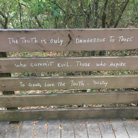 A partial view of a pedestrian walkway through the woods.  Focus of the shot are the path's side railings, consisting of 5 horizontal, parallel boards with spacing between, painted light brown and supported by vertical posts.  Upon the top three boards are written (in white chalk) "The truth is only dangerous to those who commit evil.  Those who aspire to good, love the truth.  -Tolstoy"