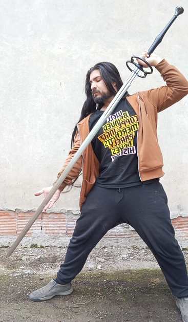 A long-haired and bearded man stands outside a building a schlachtschwert - a two-handed late medieval battle sword. The sword is nearly a foot longer than the smith is tall, and the smith is holding it diagonally with one arm raised to show the whole weapon without the blade touching the ground.
