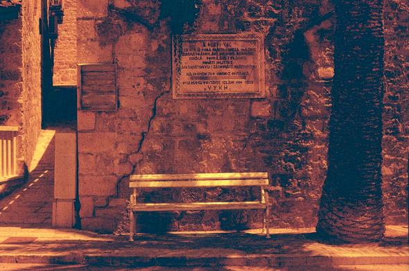 A redscale photo of a white bench next to a cracked stone brick wall and a palm tree. Above the bench there is a plaque written with an ancient greco-roman inspired font I can barely decipher,  commemorating the 700th year anniversary of Splitska in 1928